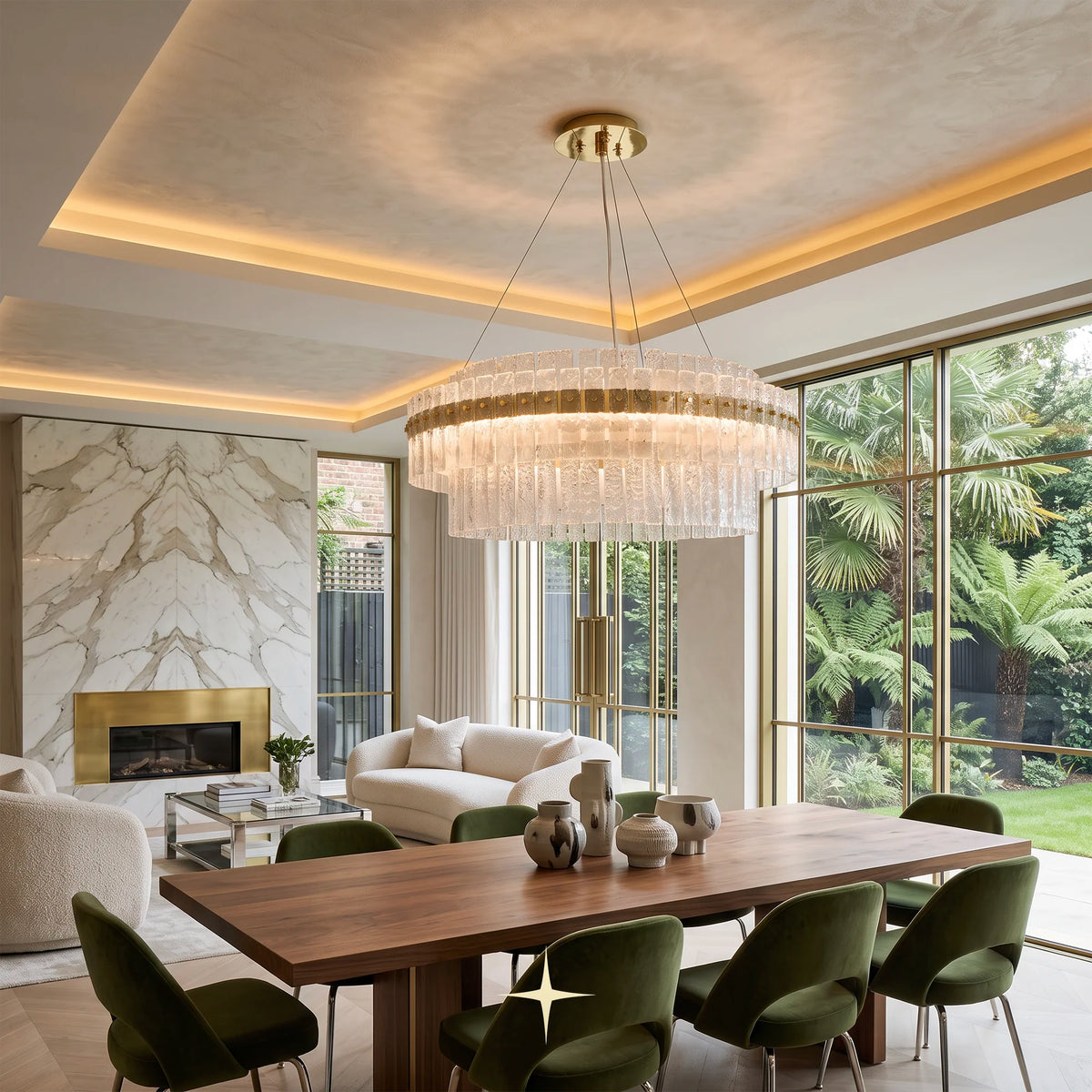 A large circular chime chandelier with a gold-toned frame and clear hanging prisms, suspended over a wooden dining table in a modern, sunlit living room with large windows and outdoor greenery.