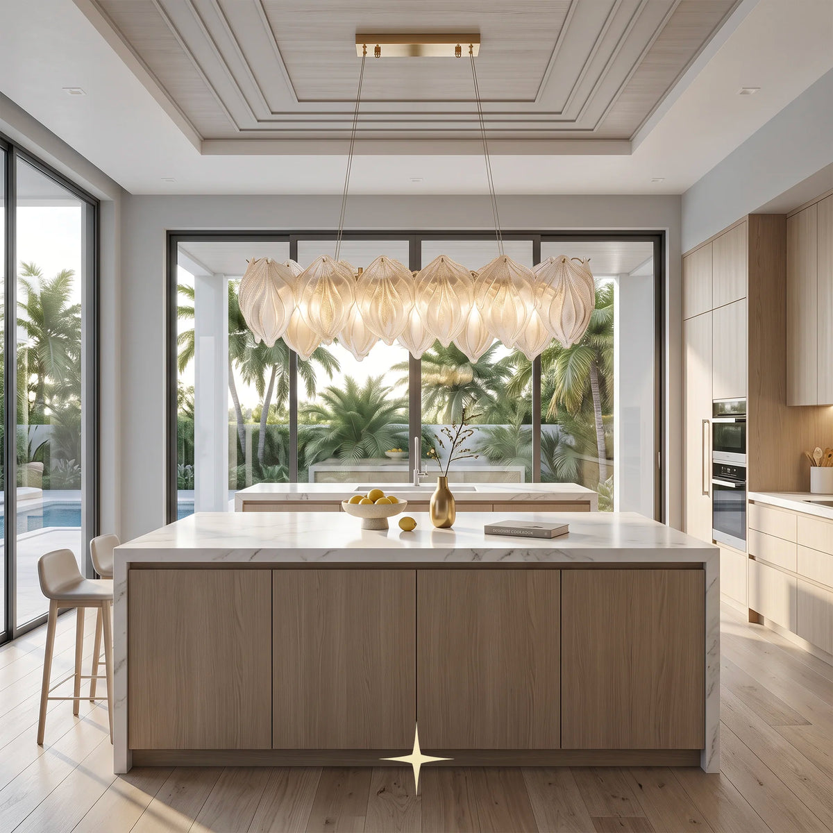 A modern kitchen island with light wood grain cabinets and a white marble countertop, under a large sculptural pendant light. The room features a high coffered ceiling, large windows with a view of palm trees, and a pool area outside.