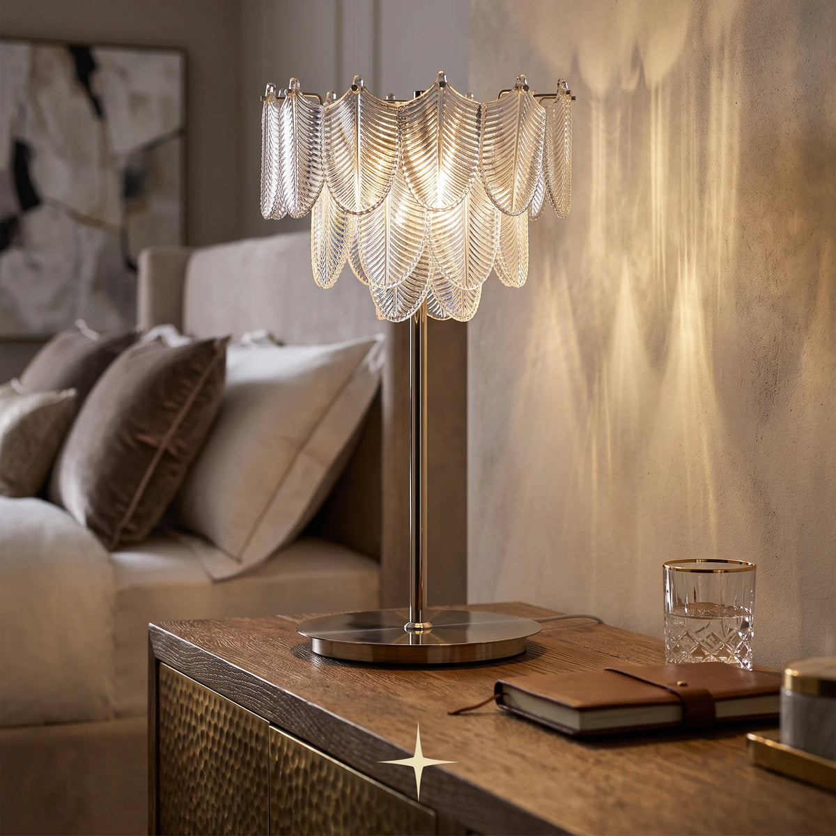 Nola Murano style glass leaf table lamp with golden nickel, scalloped clear glass shade and a brushed gold metal base, illuminated on a wooden bedside table in a warm, contemporary bedroom setting.