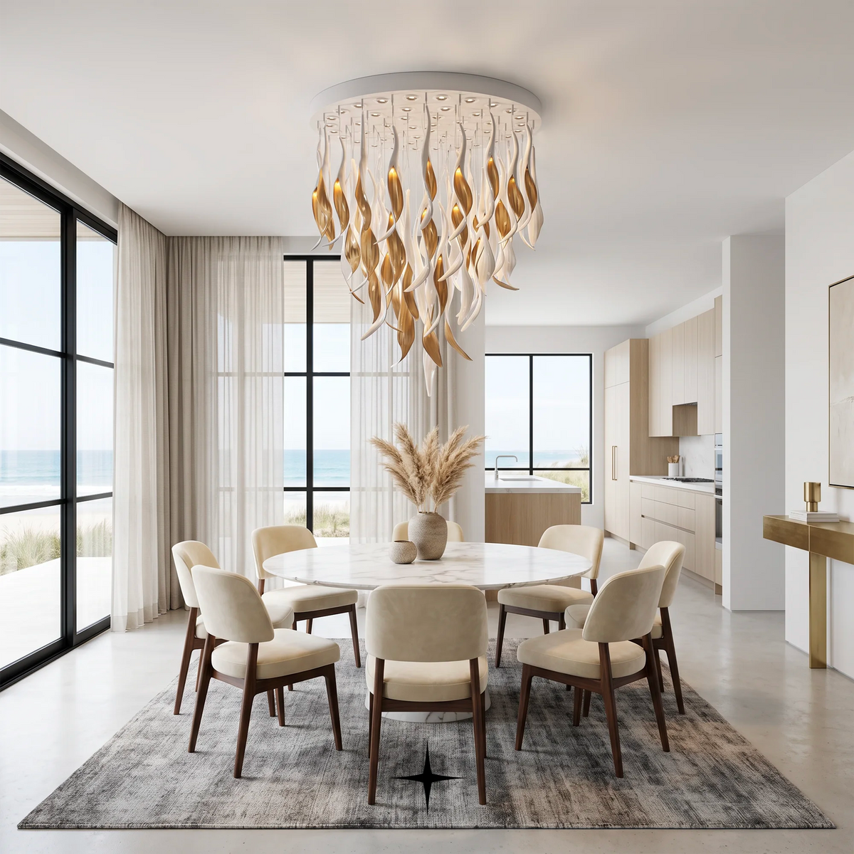 A modern dining room with a round white dining table, eight cream upholstered chairs with dark wood frames, a gray patterned area rug, and a large circular chandelier with wavy gold and white leaf-like lights hanging above. Large windows with sheer curtains look out to the ocean, and a kitchen is visible in the background.