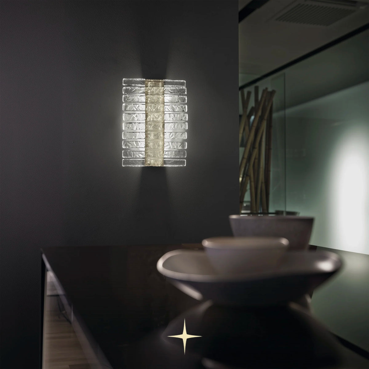 A modern rectangular wall sconce with textured clear glass panels and a central brushed gold metal strip, mounted on a dark gray wall above a table with bowls.