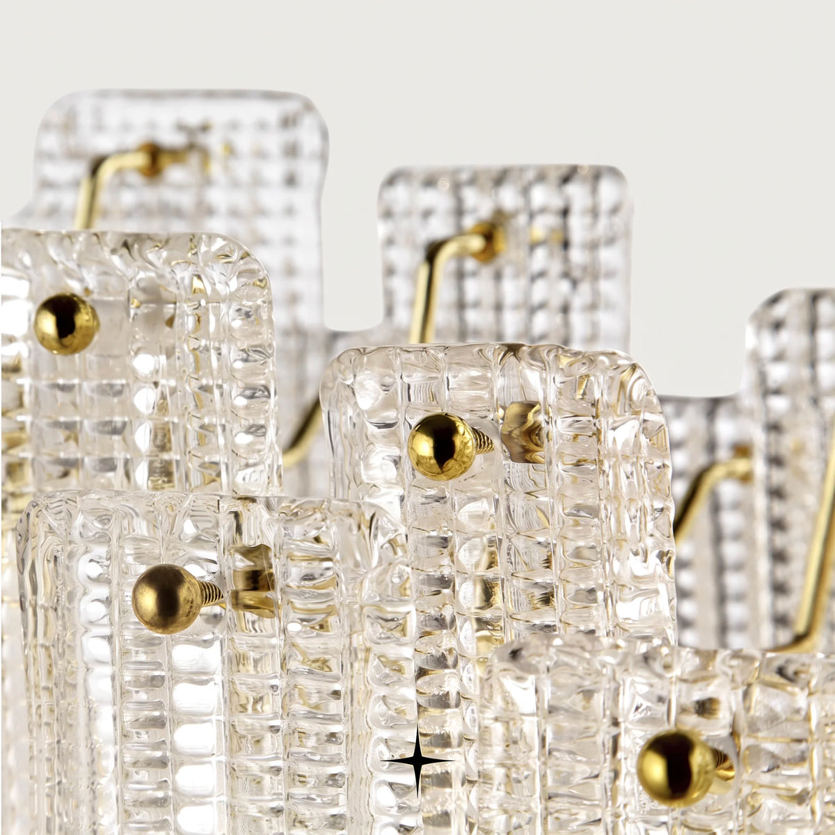 A close-up of a modern chandelier featuring clear textured glass panels with gold-finished metal accents and hooks, set against a light neutral background.