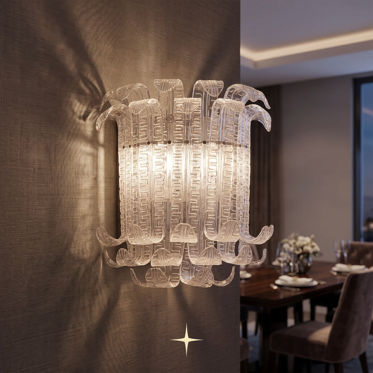 A Montreal wall sconce with sculptural, curved clear acrylic or glass arms, illuminated with a warm glow against a textured brown wall. The background shows a softly blurred dining room interior.