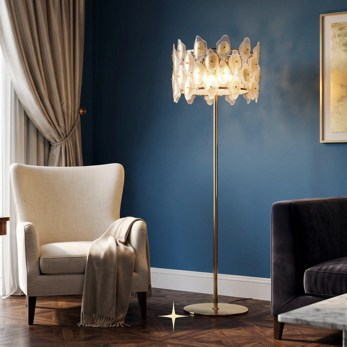 Raquel luxury floor lamp with gold and clear crystal diffusers on a polished brass with a glowing, petal-like shade and a slim gold stand, positioned in a styled living room next to a beige armchair and a dark blue wall.