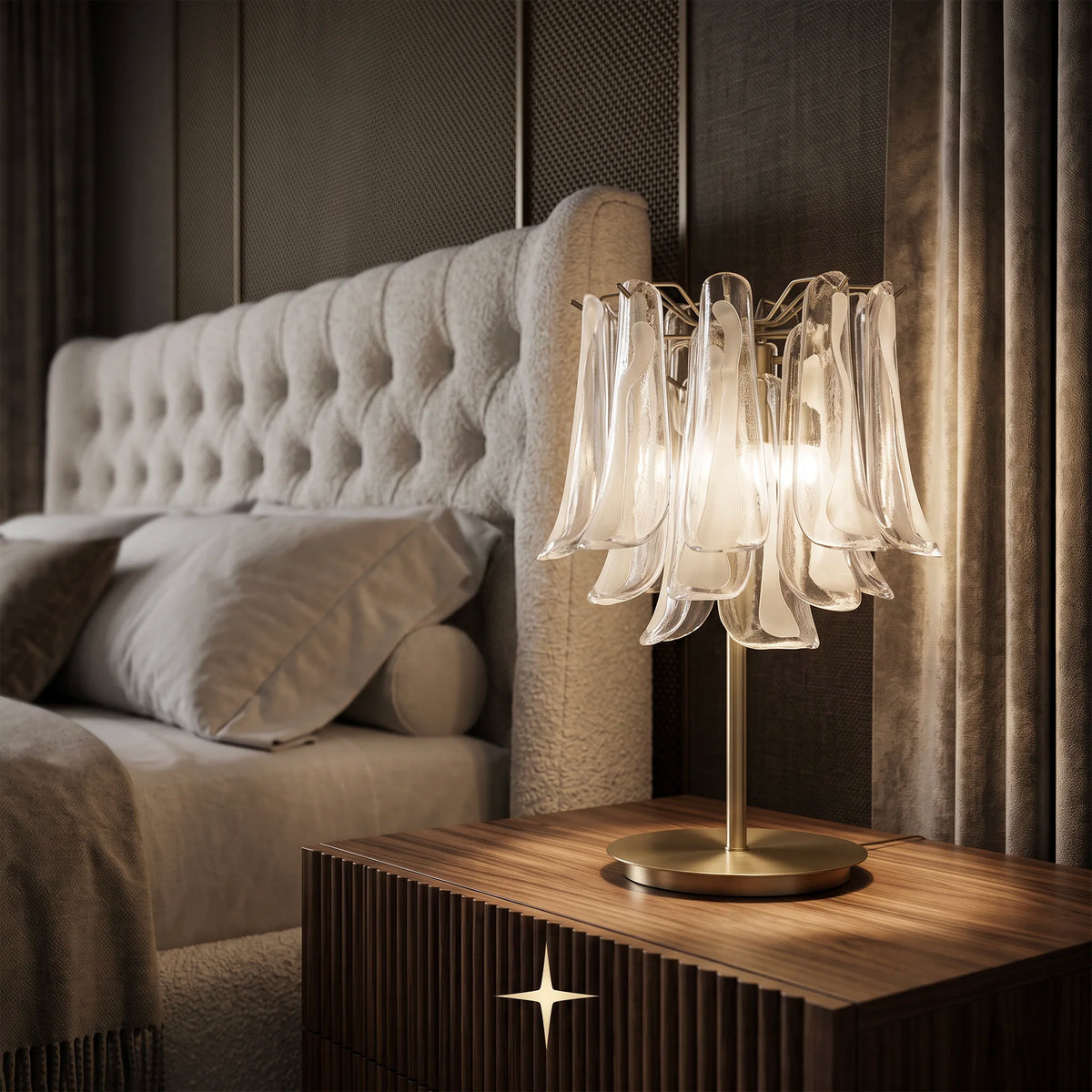 A Tranquility Murano table lamp with a warm glowing glass shade and a gold-toned metal base, placed on a wooden bedside table next to a bed in a dimly lit bedroom.