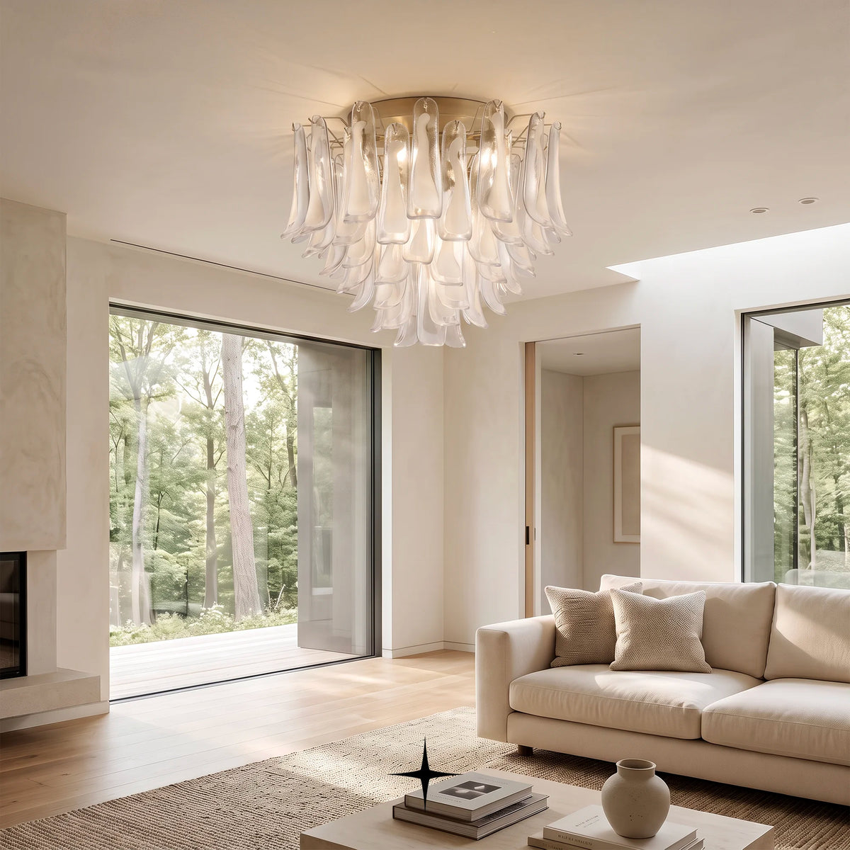 A modern chandelier with cascading translucent glass petal-like drops hangs in a bright, contemporary living room with a cream sofa, light wood floors, and large windows overlooking a wooded outdoor area.
